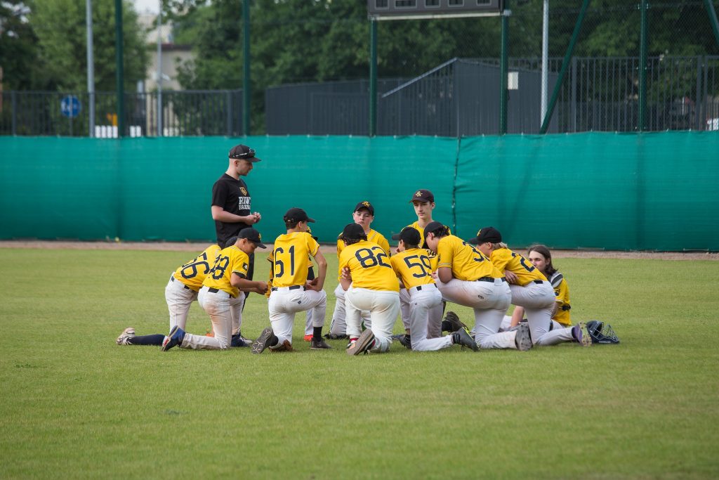 Grupa młodych baseballistów gromadzi się po meczu, rozmawiając z trenerem na boisku baseballowym w Katowicach. Zawody dla dzieci.